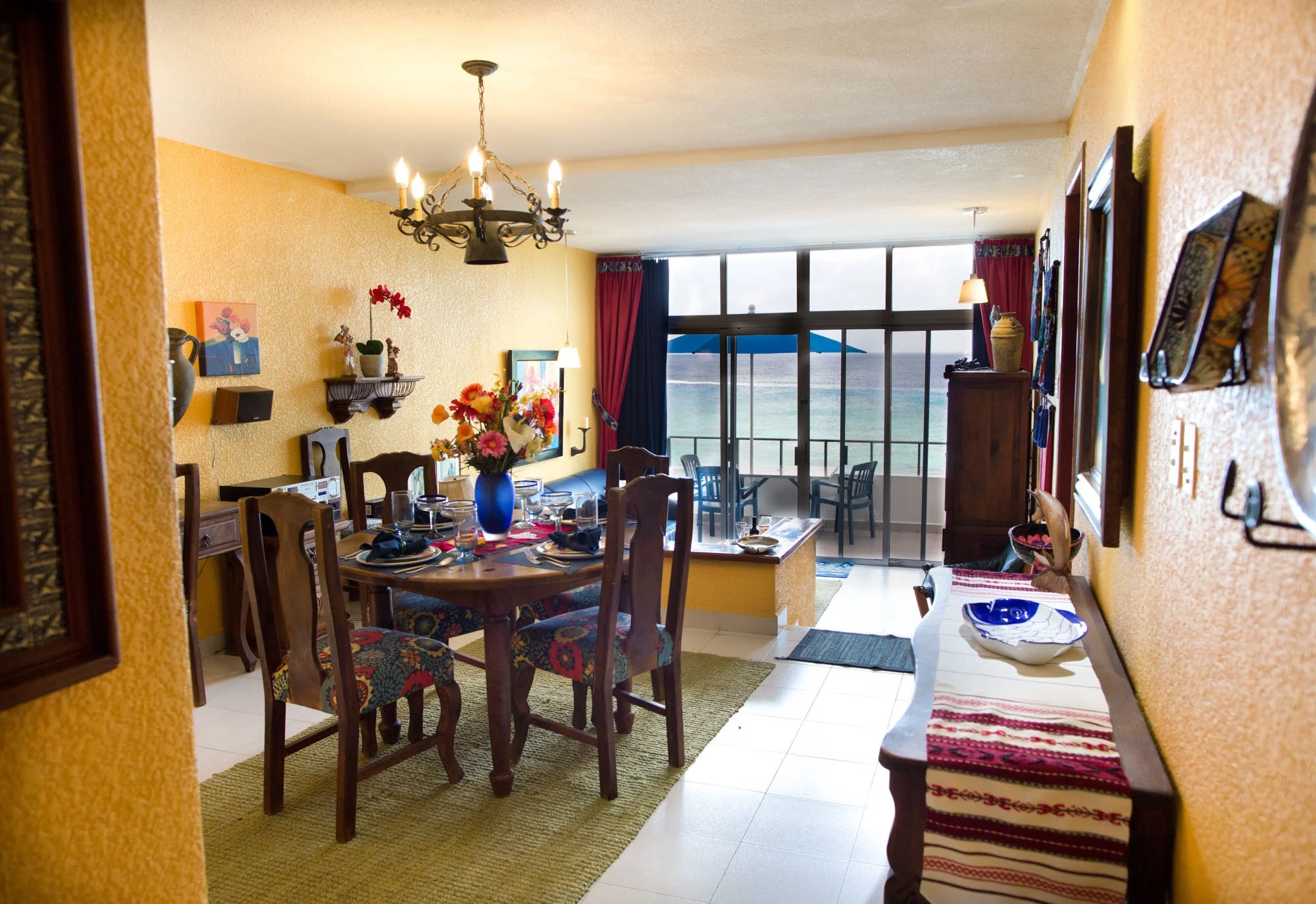 Dining room with a wooden table set for four, with a blue vase of flowers, on a green rug. Yellow textured walls, colorful chairs, and a ceiling chandelier. A sideboard with decorative items, and a view of the ocean with a balcony and patio furniture