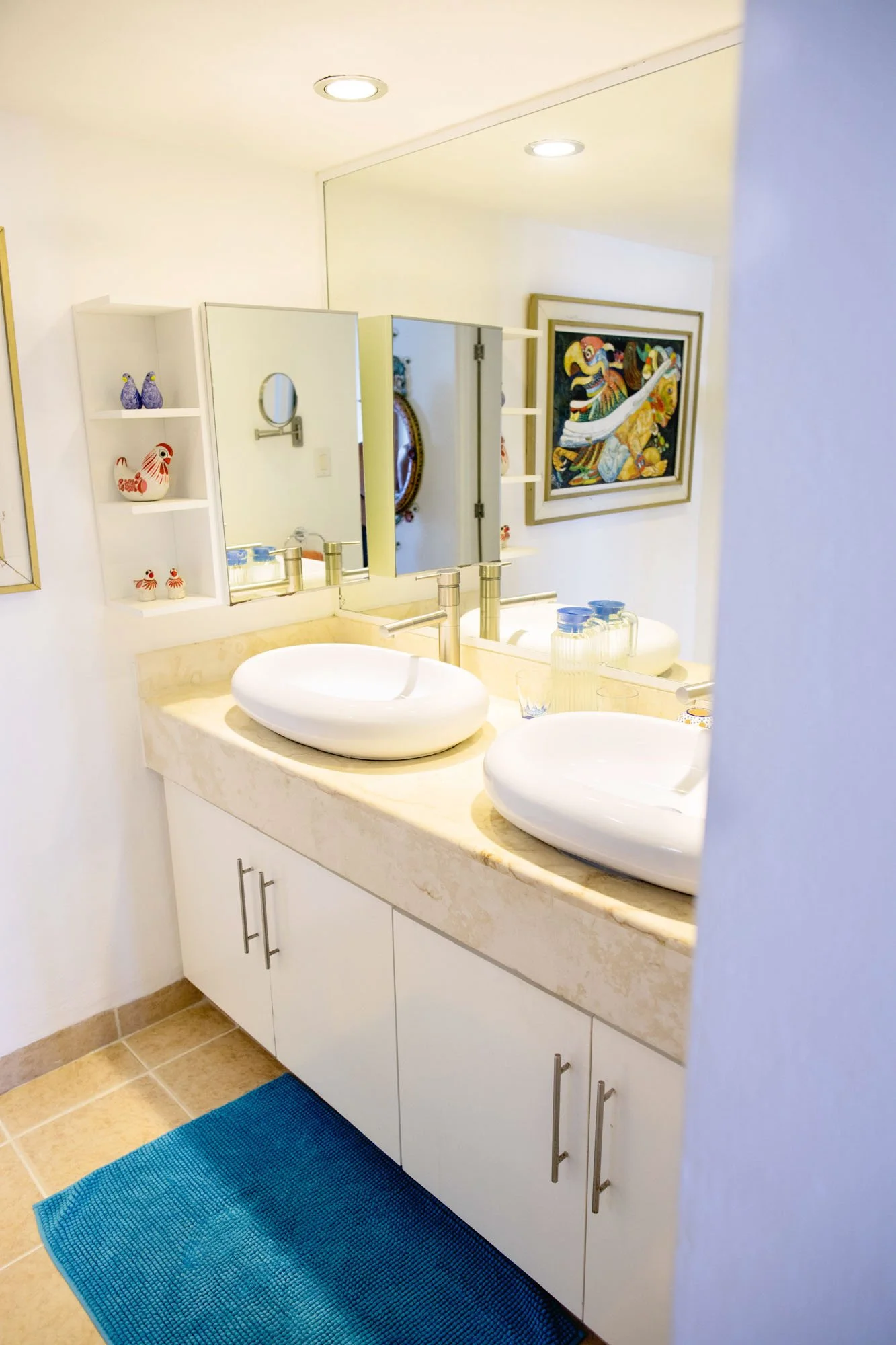Bathroom vanity with double sink, round vessel sinks, a marble countertop, cabinet below, a large mirror with side shelves displaying colorful decorative items, and framed artwork on the wall.