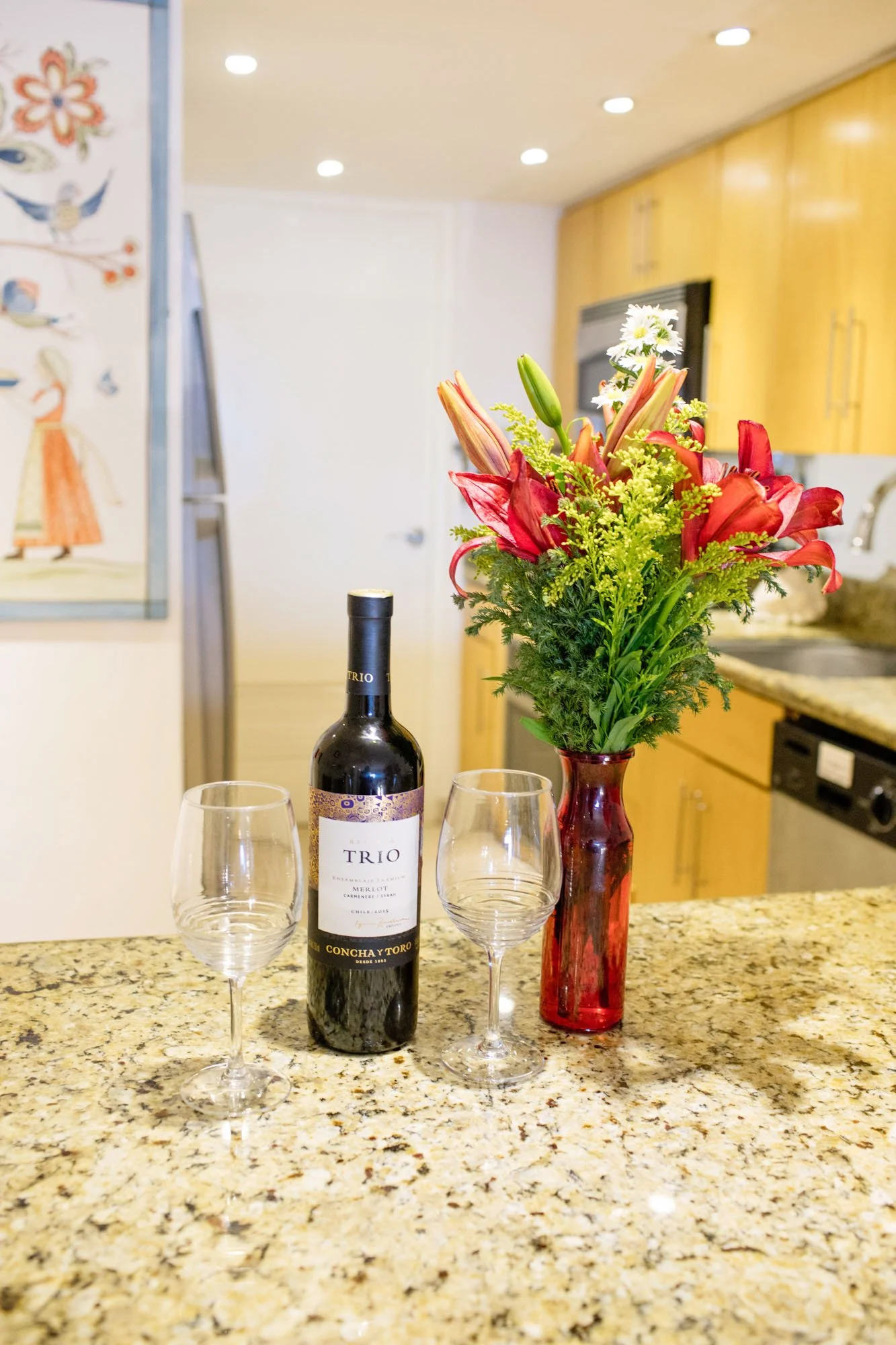 A bottle of red wine, two empty wine glasses, and a red vase with a colorful flower arrangement on a kitchen granite countertop.