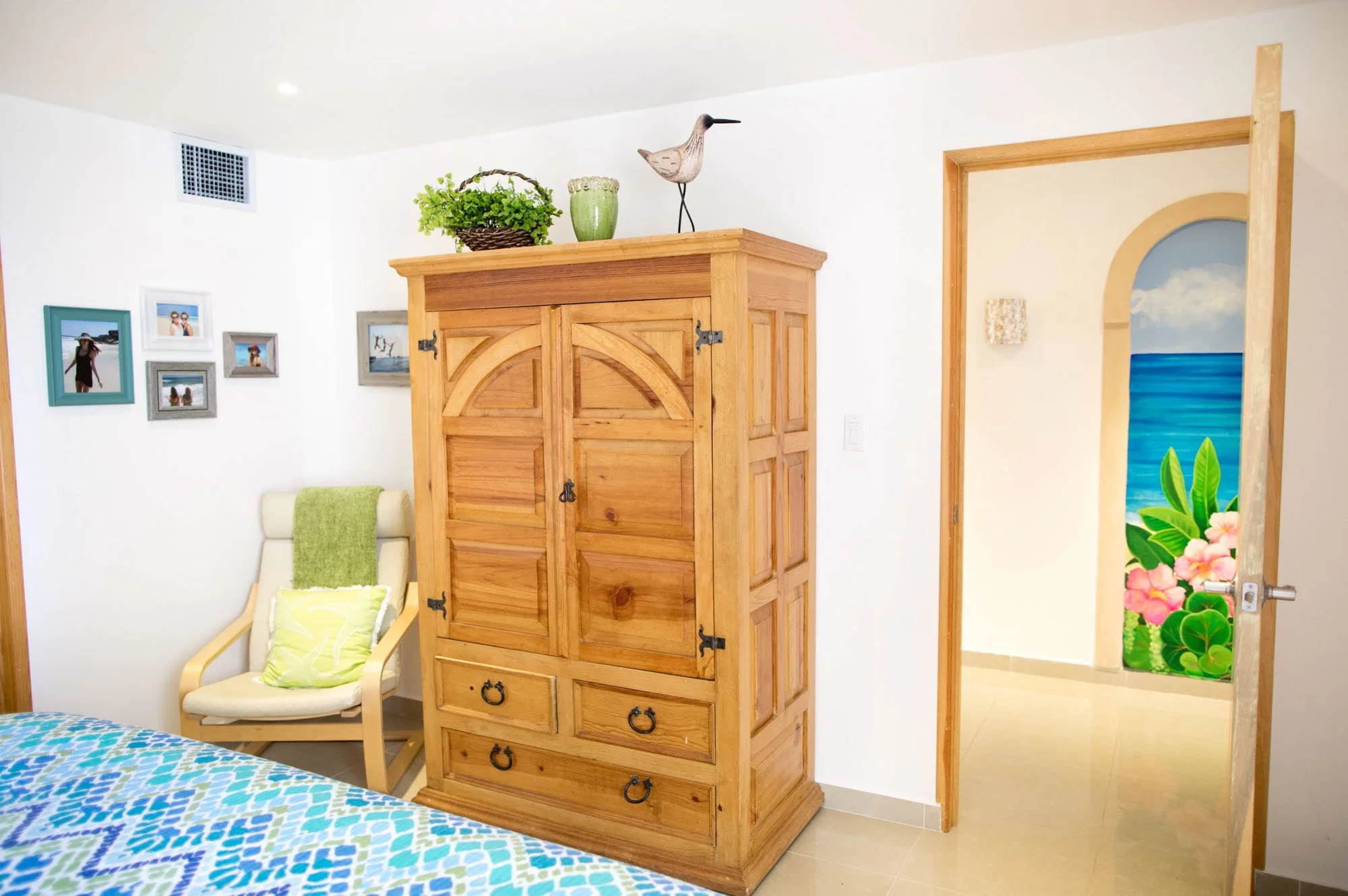 Bedroom with wooden wardrobe, armchair with green pillow, photo frames on wall, decorative items on wardrobe, and a door with a ocean and floral mural.