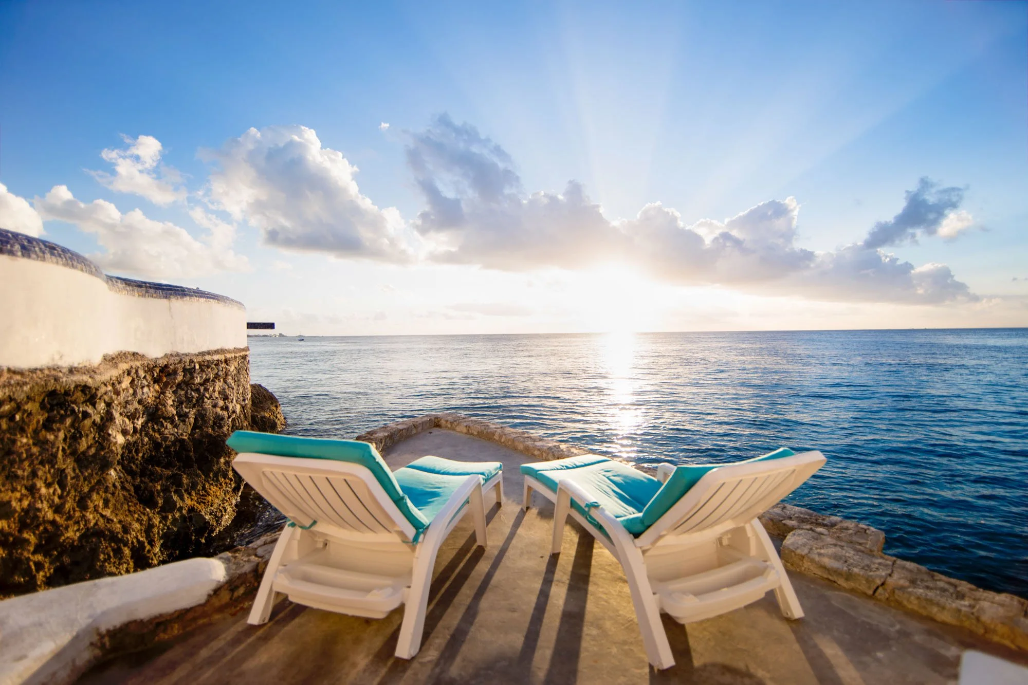 Two lounge chairs with blue cushions facing the ocean, with the sun setting on the horizon and clouds in the sky at a Cozumel vacation rental.