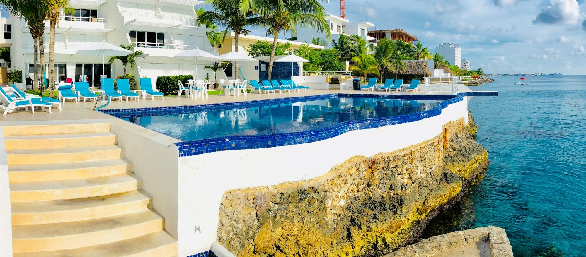 Swimming pool with blue tiles on a rocky base, surrounded by lounge chairs and umbrellas, overlooking the ocean in a tropical resort.