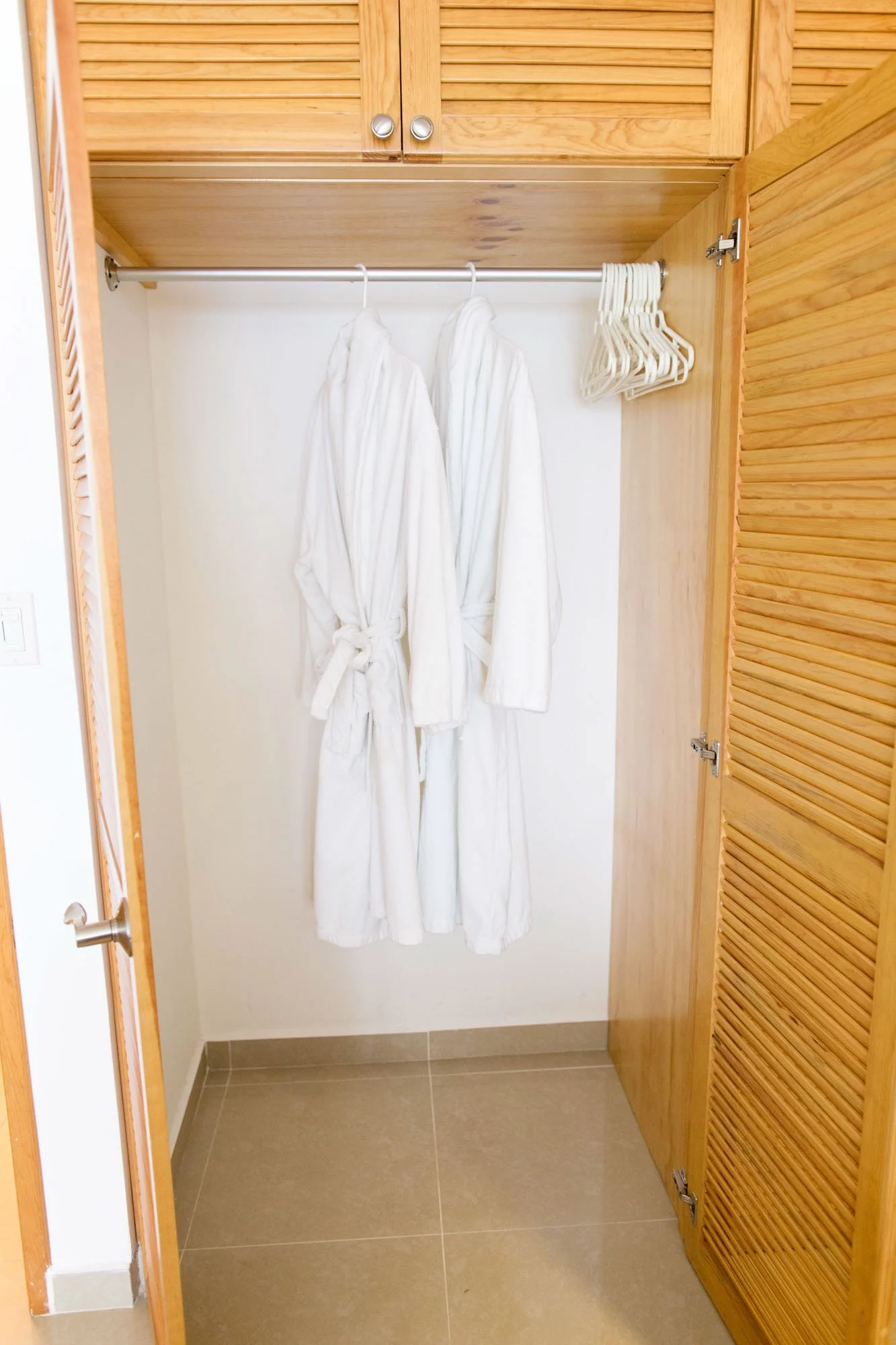 Open wooden closet with two white bathrobes hanging on hanger and several empty white hangers on side.