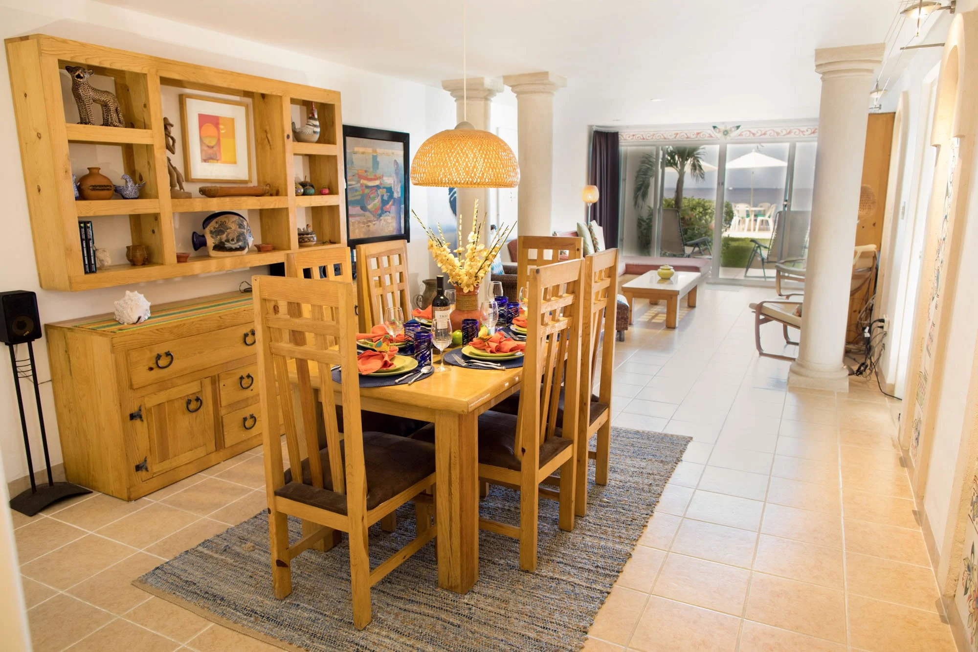 Dining area with a wooden table set for six, a large hanging light, and decorative items on a shelf on the wall at a Cozumel vacation rental.