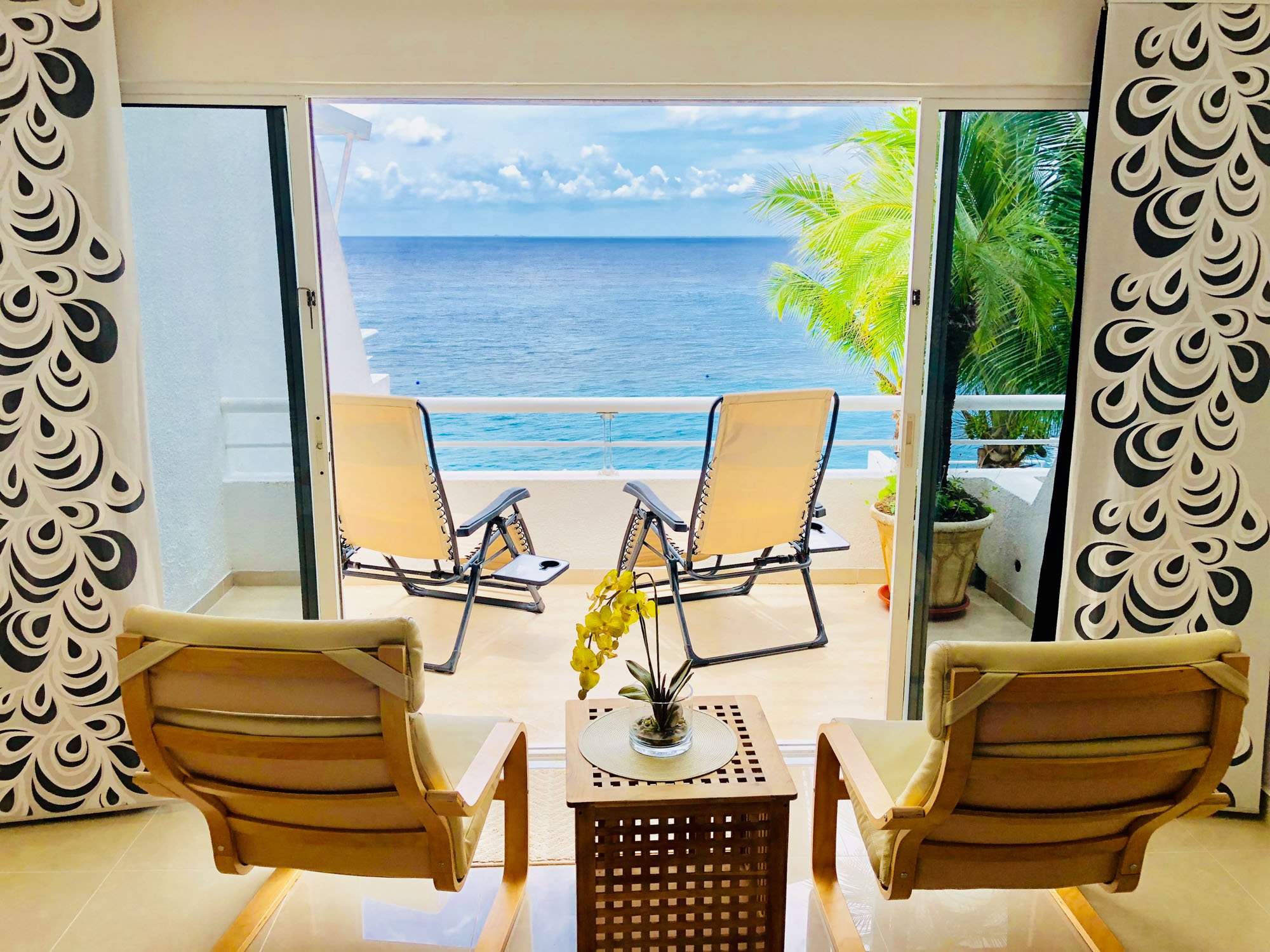 Living room with two armchairs facing a balcony with two lounge chairs overlooking the ocean, with green palm trees and a cloudy sky.