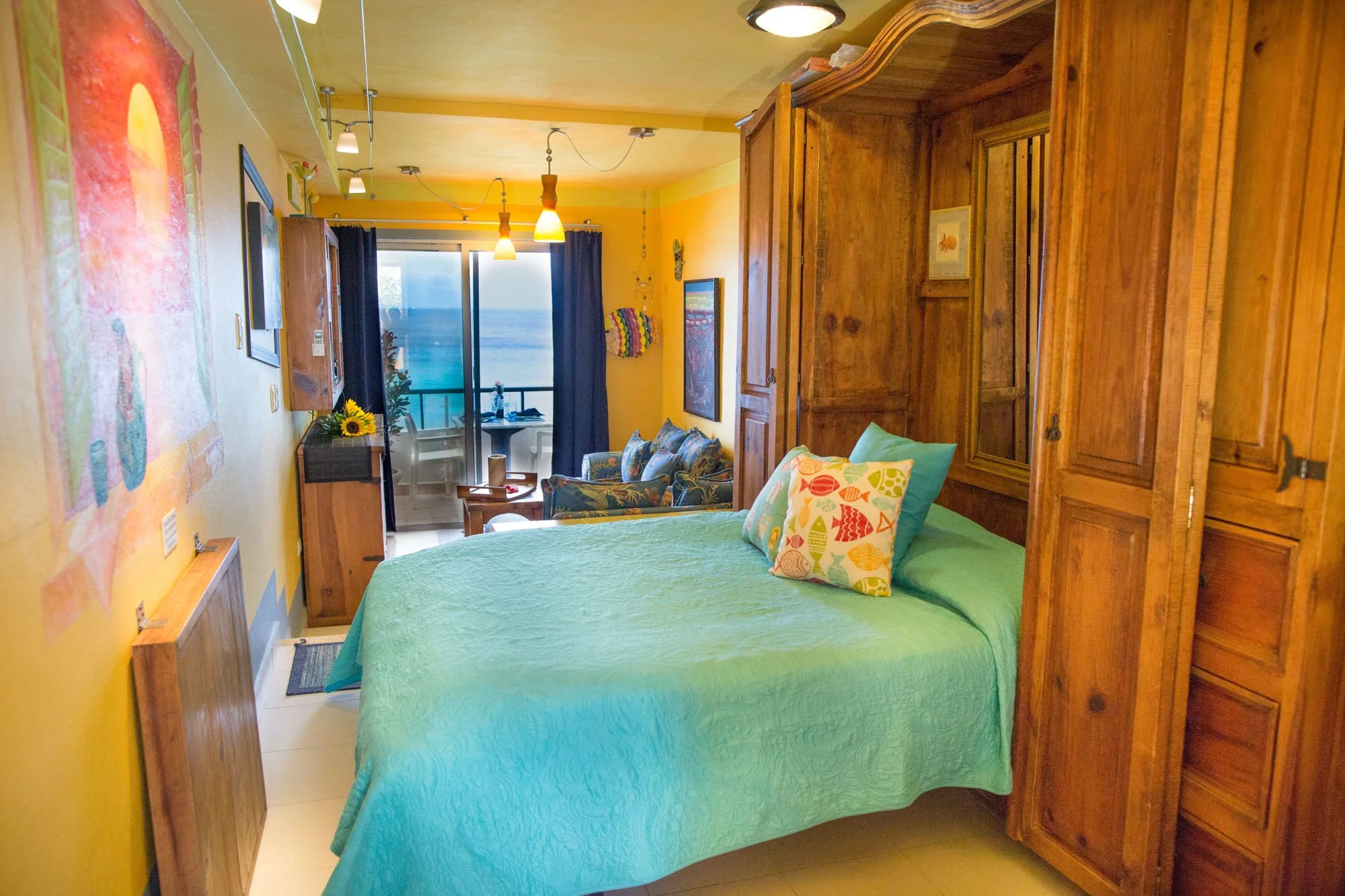 Beachfront hotel room with a bed, colorful pillows, wooden wardrobe, small sitting area, and a balcony with ocean view at a Cozumel vacation rental.