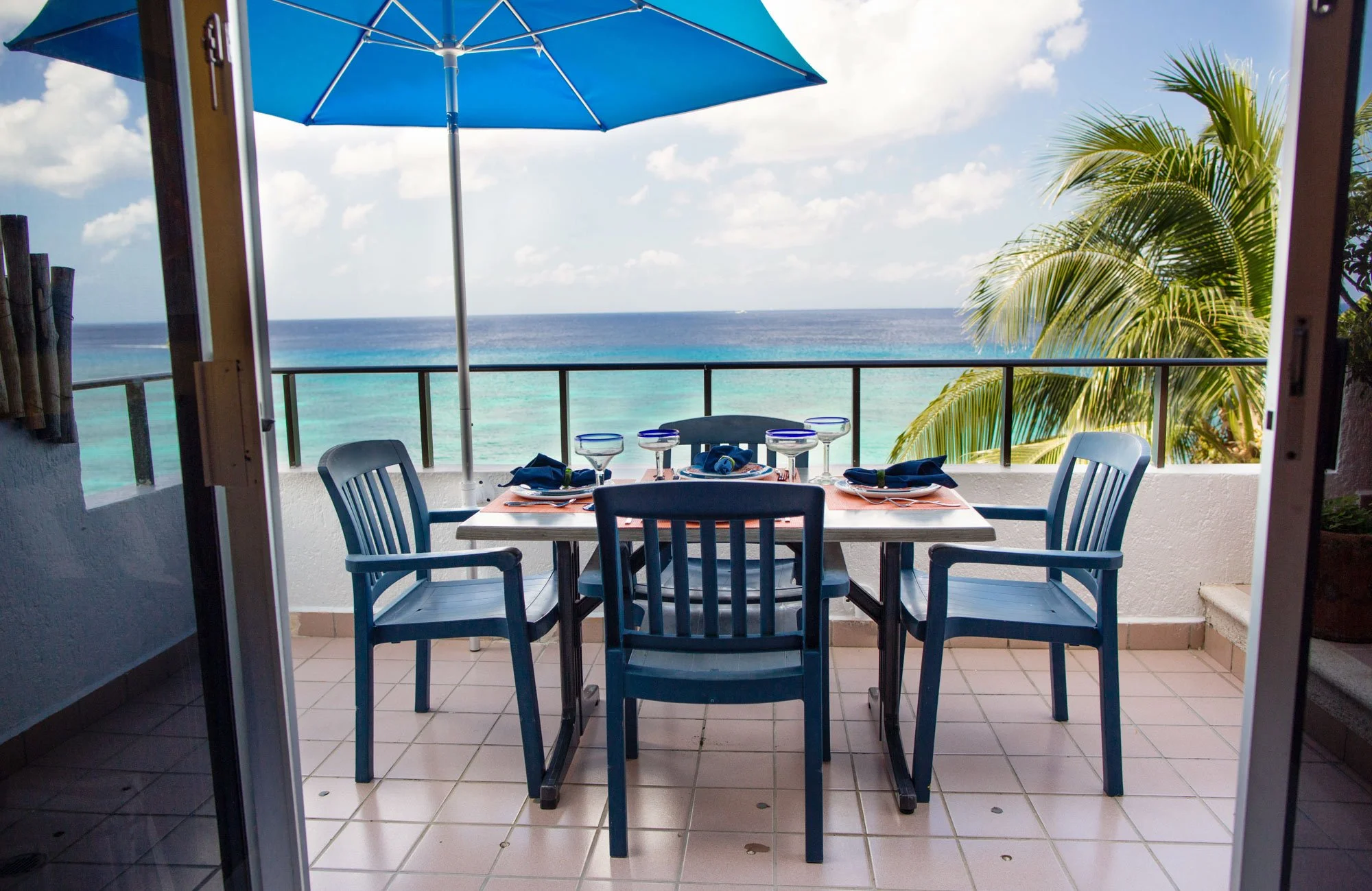 Cozumel vacation rental dining table set for four with blue chairs, glassware, and napkins on a balcony overlooking the ocean, with a blue umbrella and a palm tree.