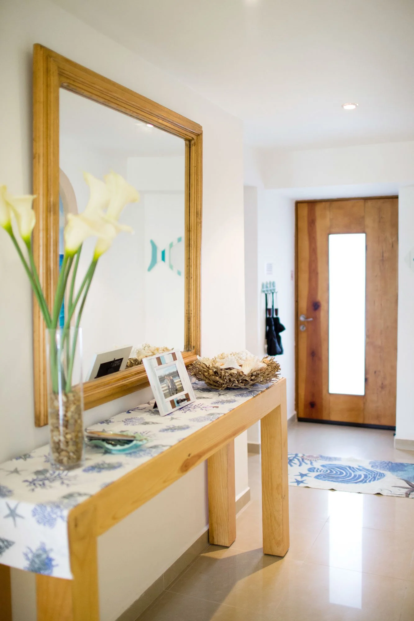 Entryway with wooden front door featuring a vertical glass panel, a hallway table with decorative items, a large mirror with a wooden frame, and a woven rug with blue seashell and starfish patterns on the floor.