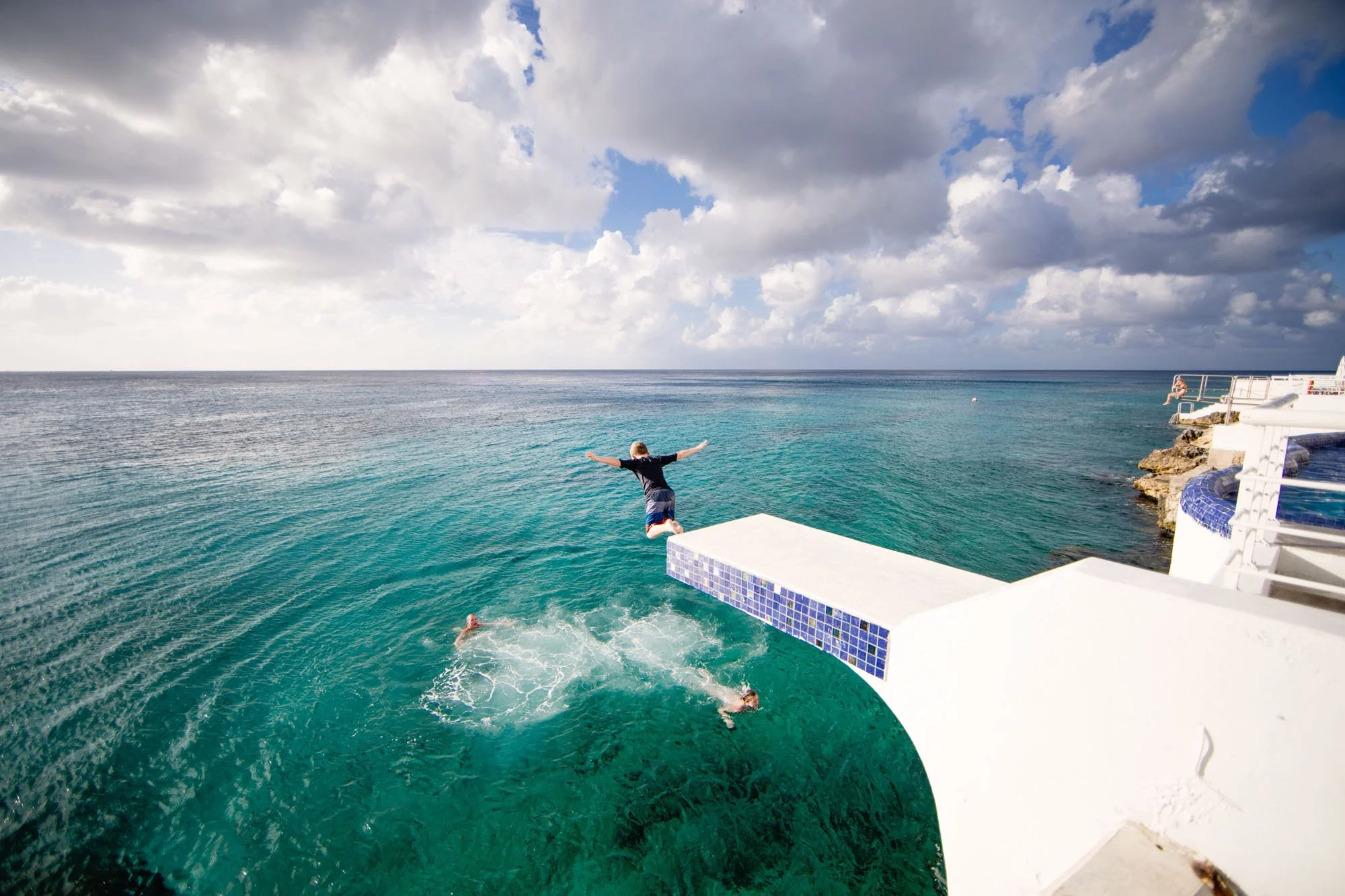 Person jumping off a diving board into the ocean under a partly cloudy sky.