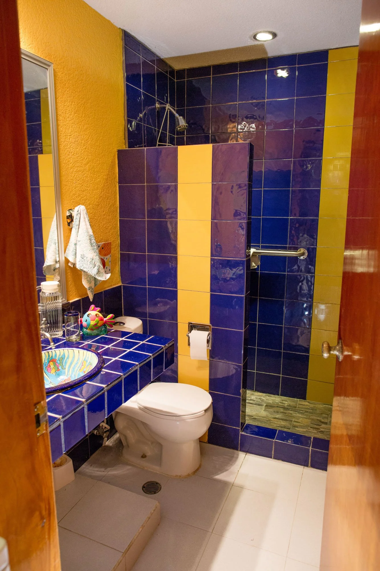 A colorful bathroom with blue, yellow, and purple tiles, a small sink with a bowl-shaped basin, a toilet, and a walk-in shower with a grab bar.
