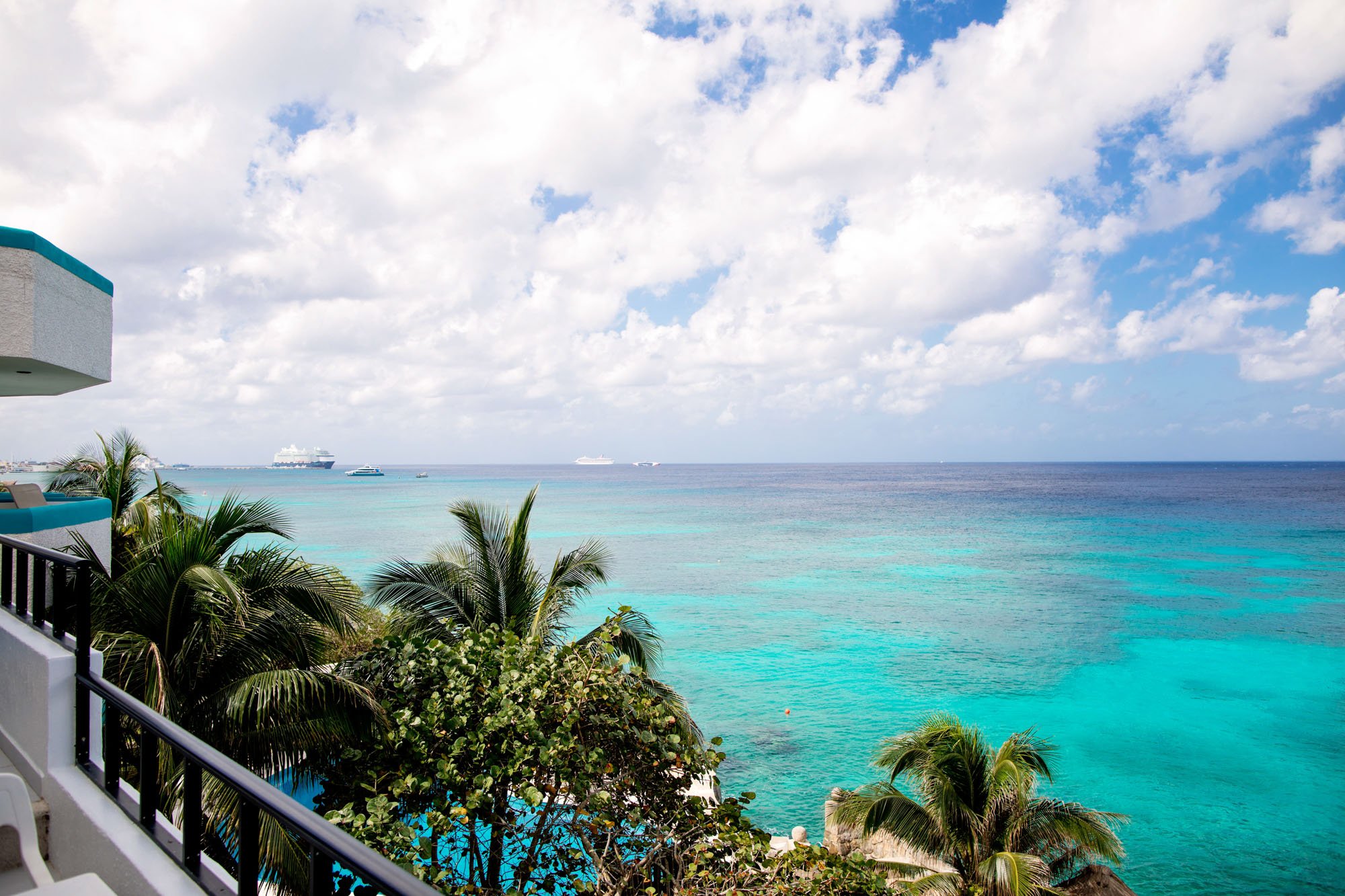 Balcony view of turquoise blue ocean with cruise ships sailing under partly cloudy sky, surrounded by green palm trees and tropical plants.