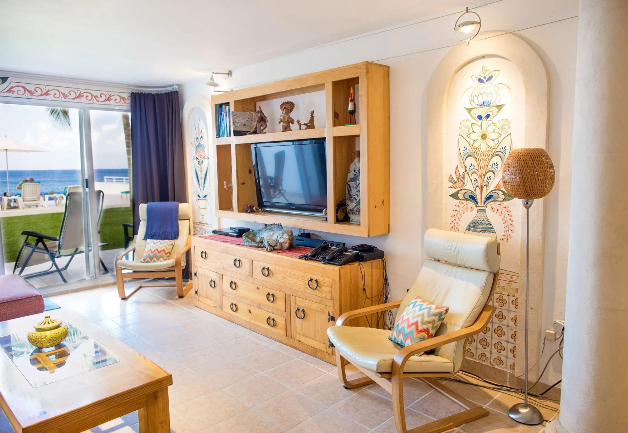Living room with ocean view, featuring chairs with colorful pillows, a wooden TV stand with multiple drawers, a flat-screen TV, decorative items, and sliding glass door leading to a balcony with outdoor seating and a view of the ocean.