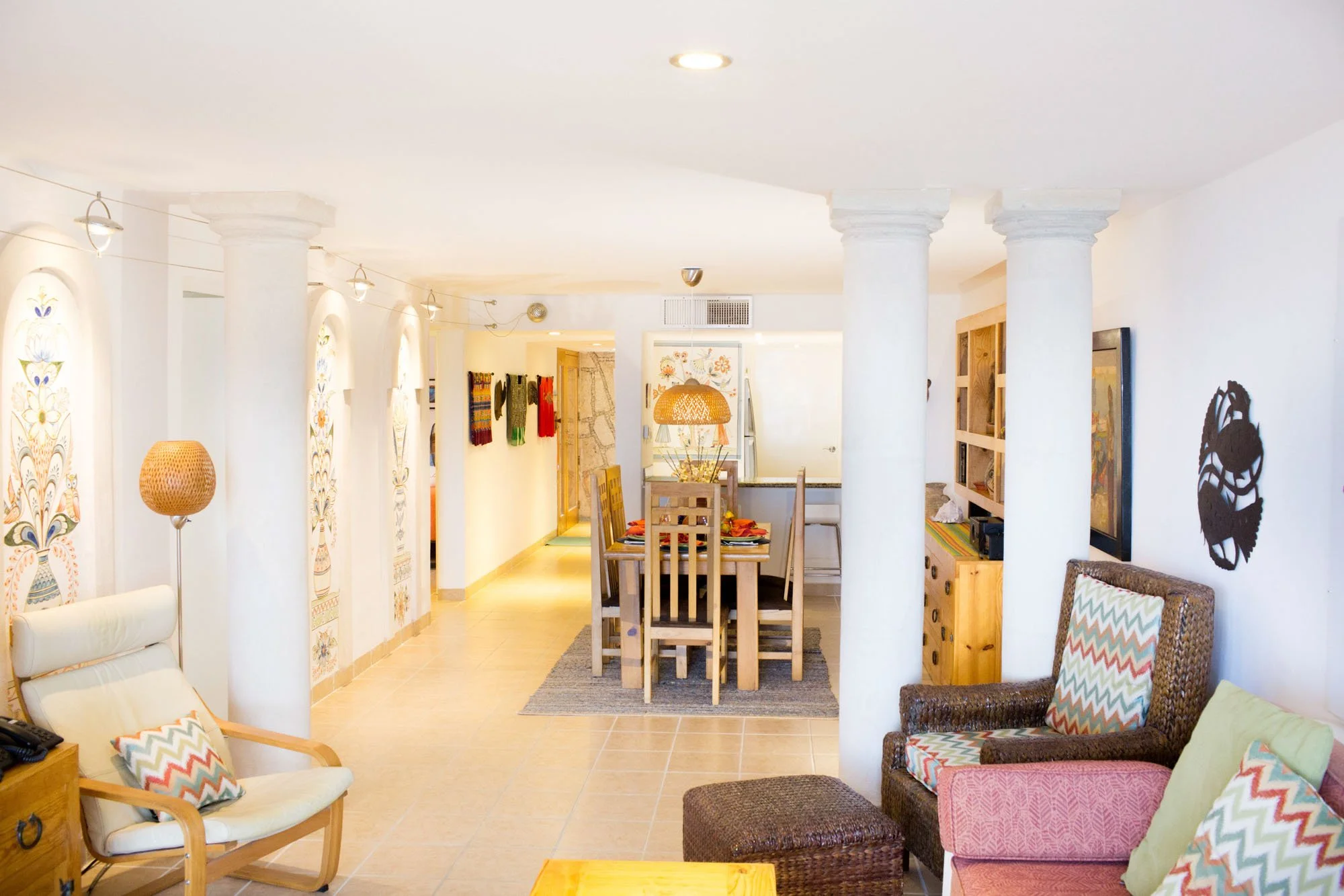 Living room with white walls, tiled floor, and wooden furniture. Includes armchairs, a wicker chair with colorful cushions, a wooden dining table with chairs, decorative wall art, and a bookshelf.