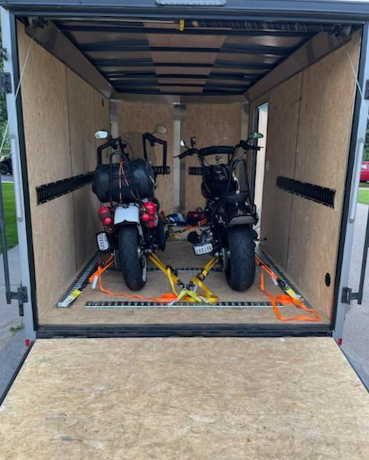 Two motorcycles inside an empty enclosed trailer with wood paneling, secured with orange straps.