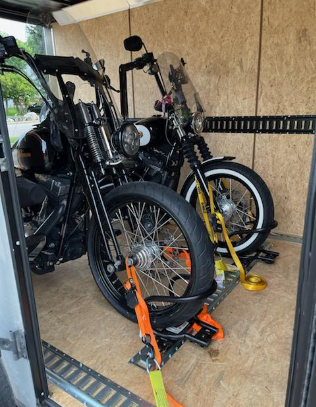 Two black motorcycles secured in a trailer with orange straps, with a yellow bicycle also secured, all parked on a wooden floor.