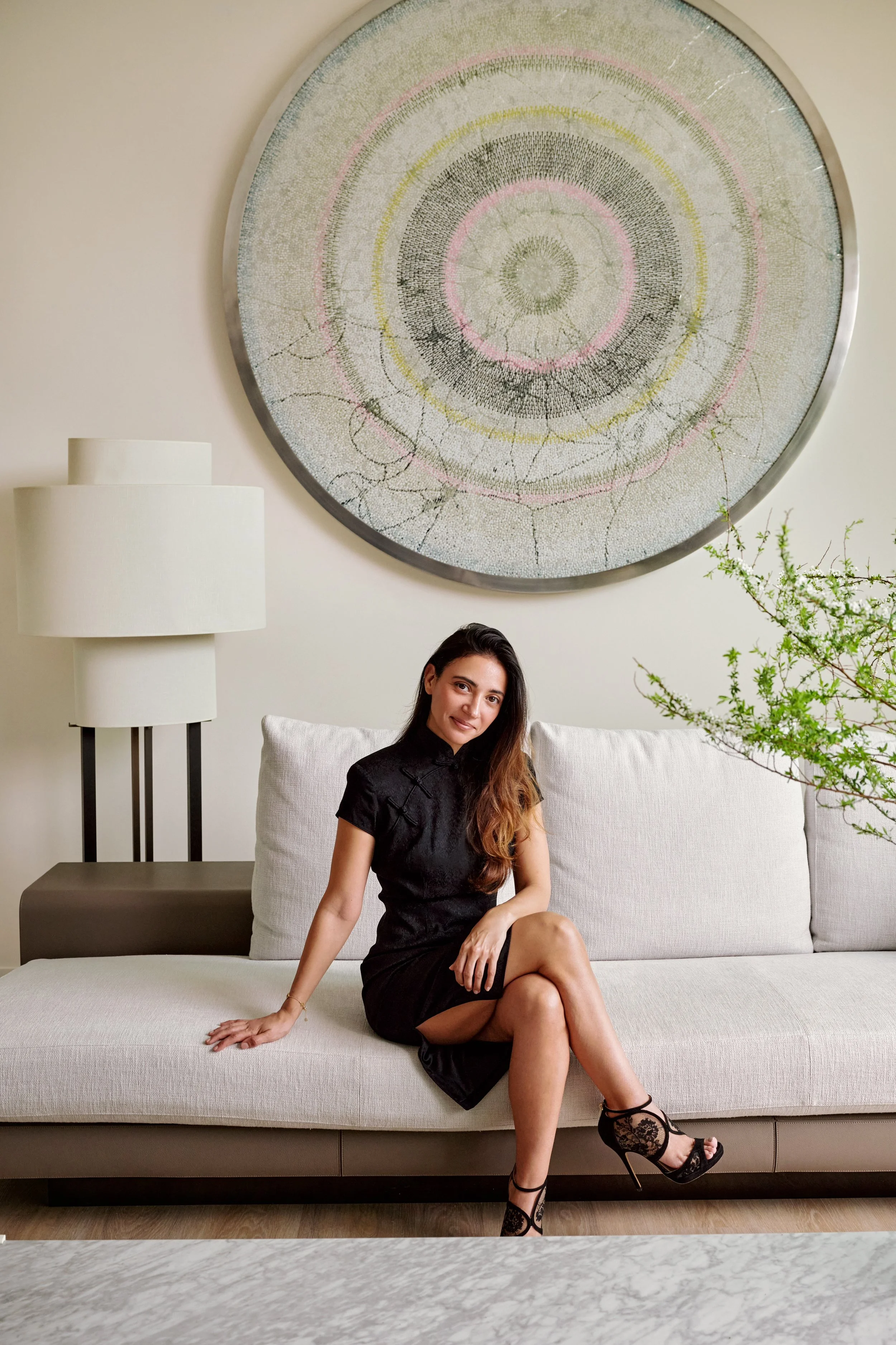 A woman sitting on a light-colored sofa in a modern living room with a large circular artwork on the wall behind her, a white lamp to her left, and a green plant to her right.