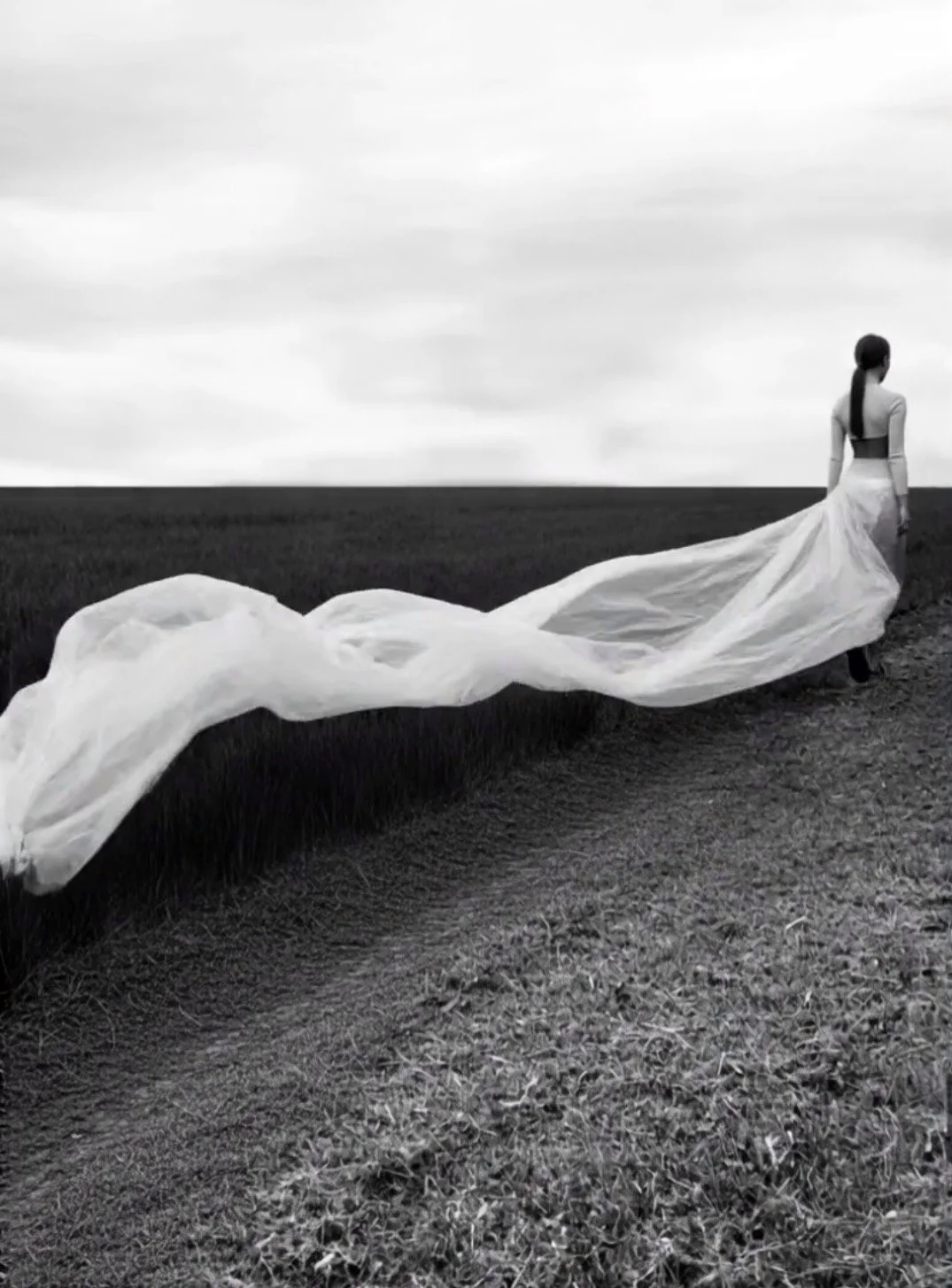 Black and white photo of a woman in a long dress walking on a field, with a long train flowing behind her.