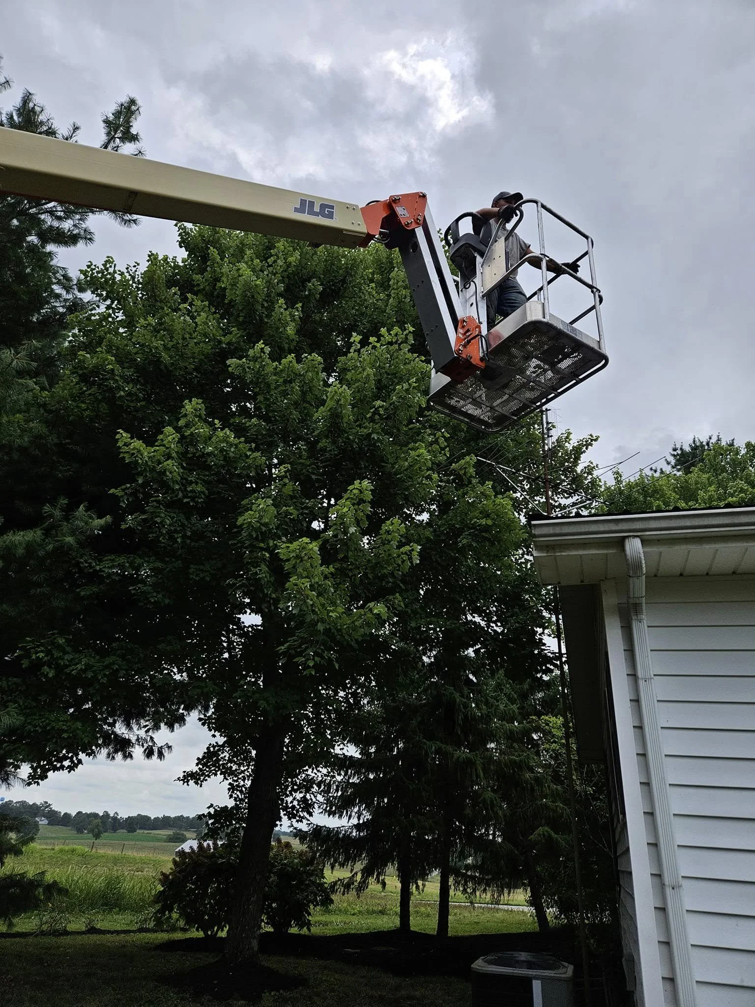 A man in a cherry picker lift working on an antenna on a house roof in a backyard with trees and a cloudy sky.