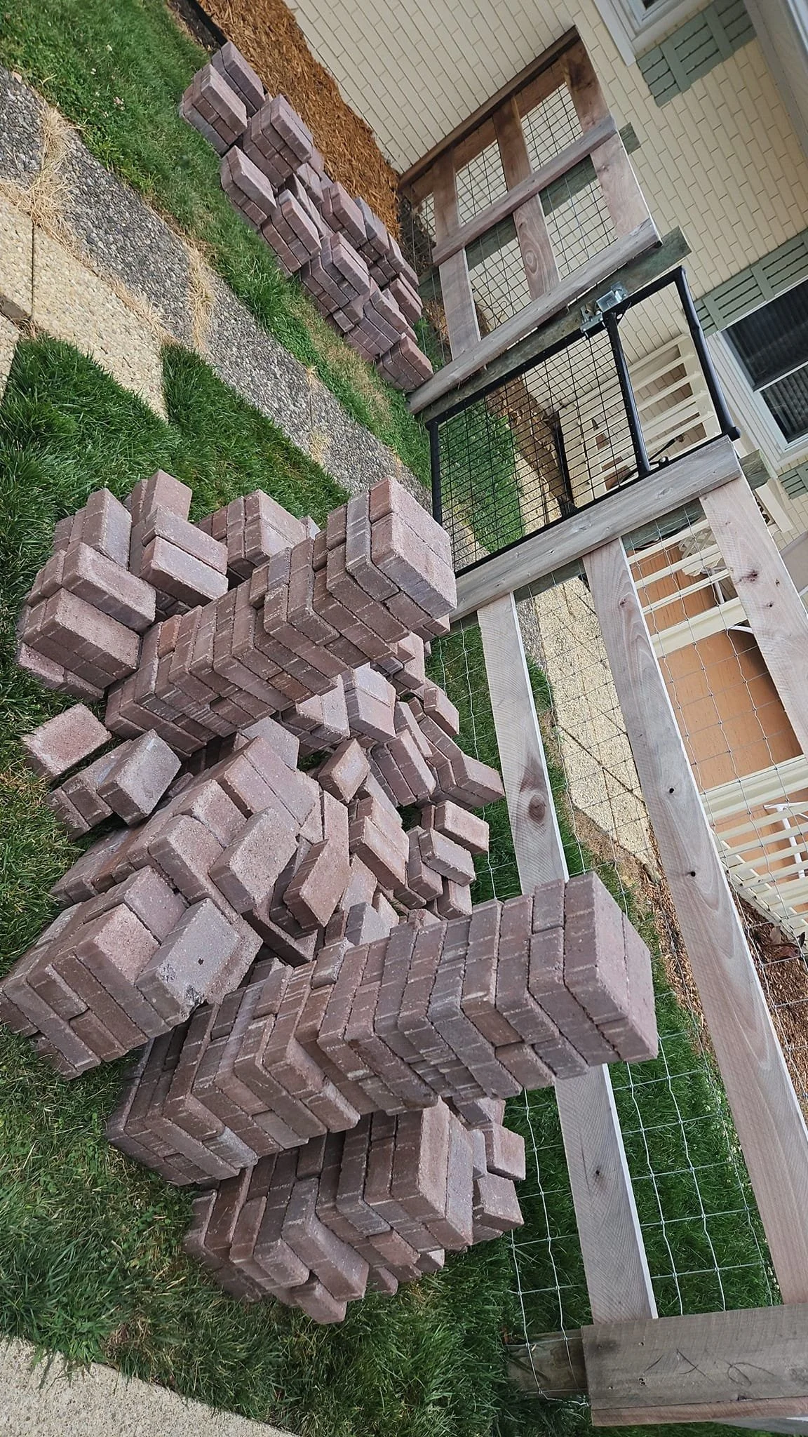 Piles of bricks stacked on grass and sidewalk near a wooden and wire fence, with a house in the background.