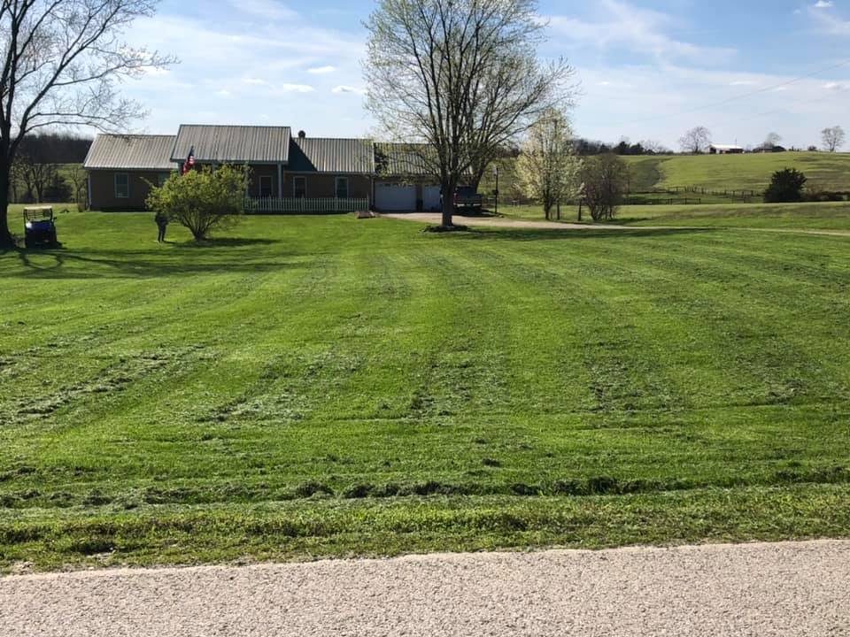 A rural house with a large lawn and trees, with a road in the foreground and rolling hills in the background on a sunny day.