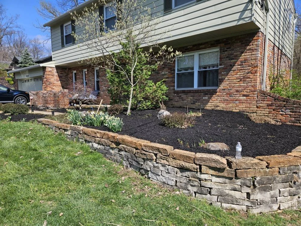 Front yard landscaping with freshly mulched flower bed, young green plants, a small tree, and a stone retaining wall in front of a house with brick and siding exterior.