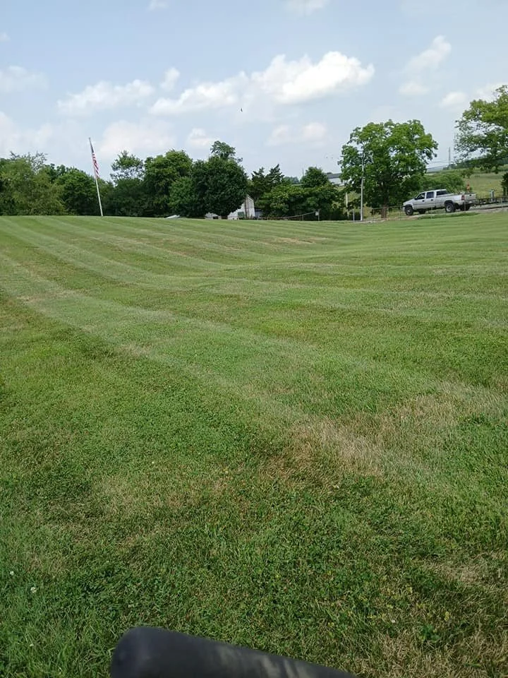 A grassy field with a slight incline, a flagpole with an American flag, trees, a white building, and a parked truck on a sunny day with partly cloudy skies.