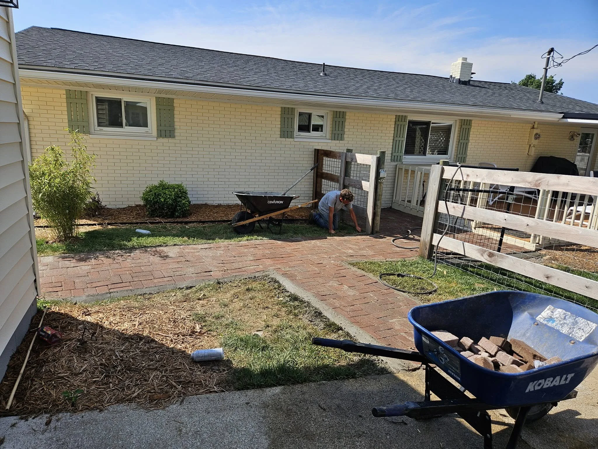 A person working on laying bricks in a backyard with a partially finished brick pathway. There is a blue wheelbarrow filled with bricks, a garden bed with mulch and some small bushes, and a fenced area. The house in the background has yellow brick walls, green shutters, and three small windows.