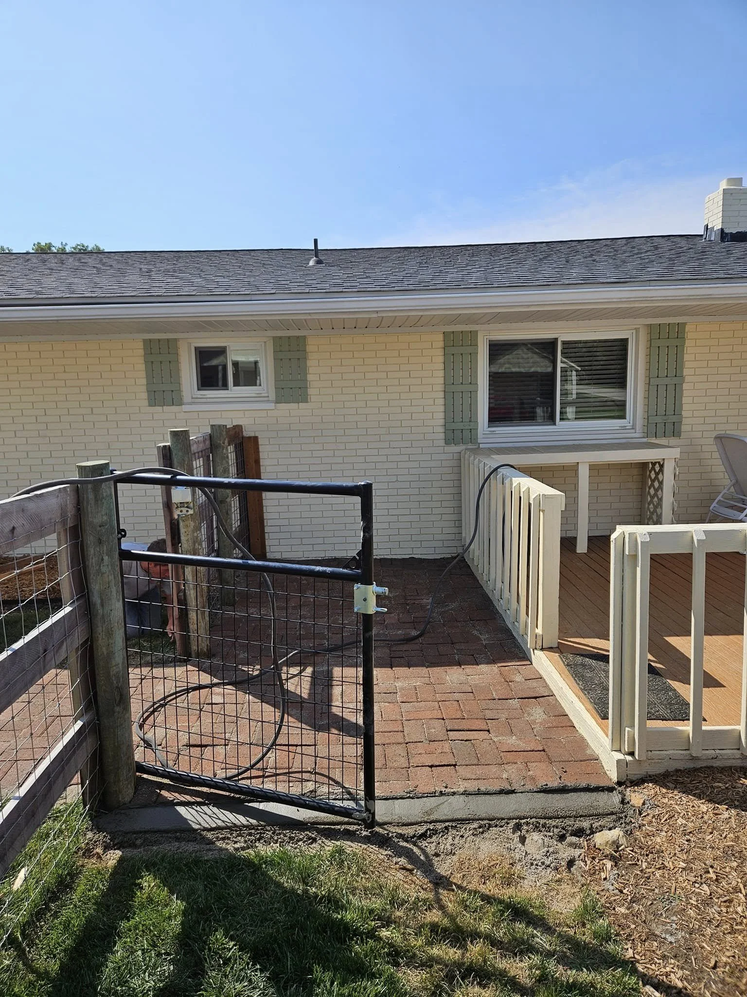 Backyard with brick patio, small white deck with railing, and a black metal gate. House has white brick exterior with green shutters and a window. Clear blue sky.