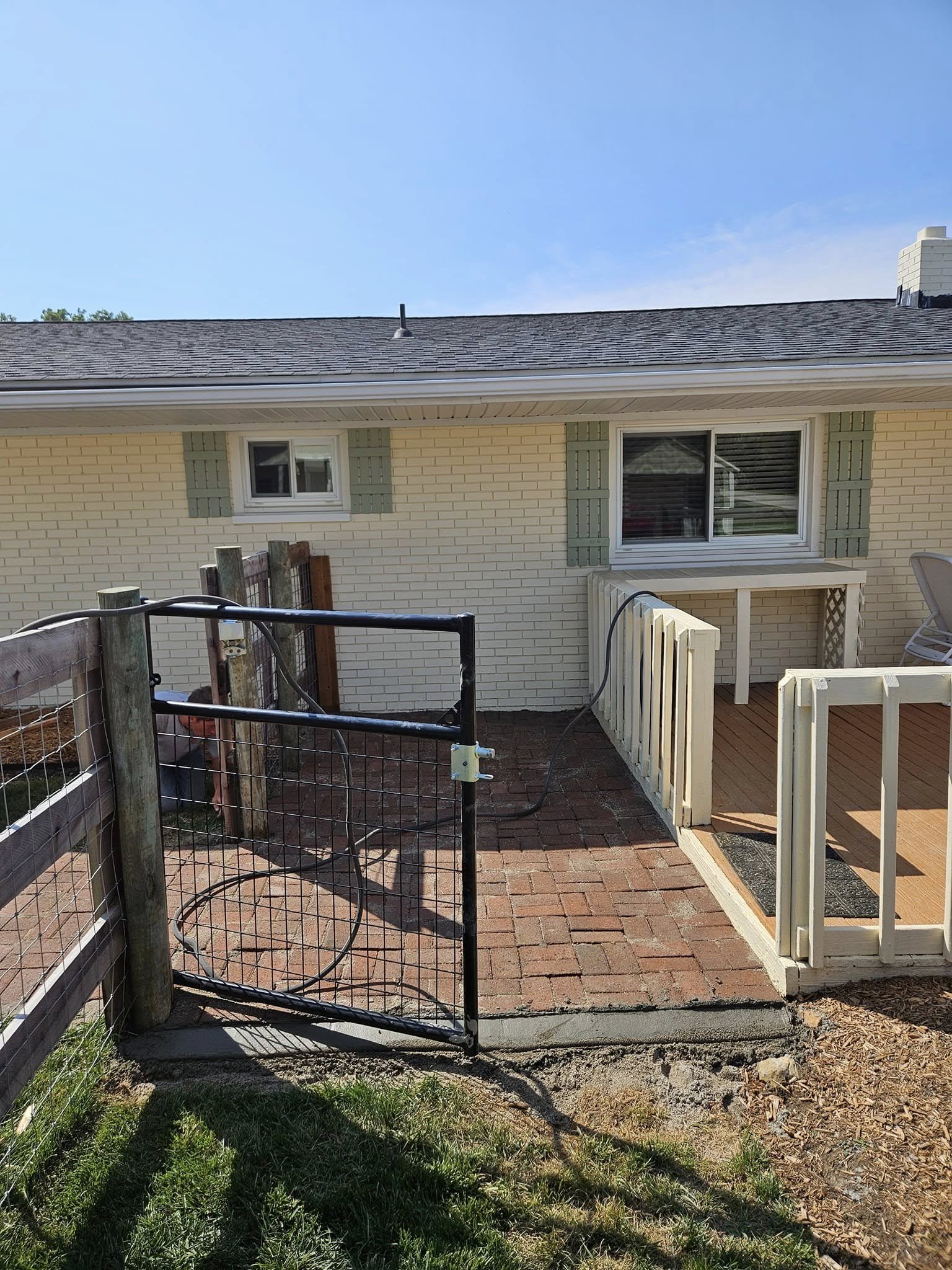 Backyard with a small brick patio, a white wooden fence, a black metal gate, and a raised wooden deck with a white railing. House with white brick walls, small windows, and green shutters.