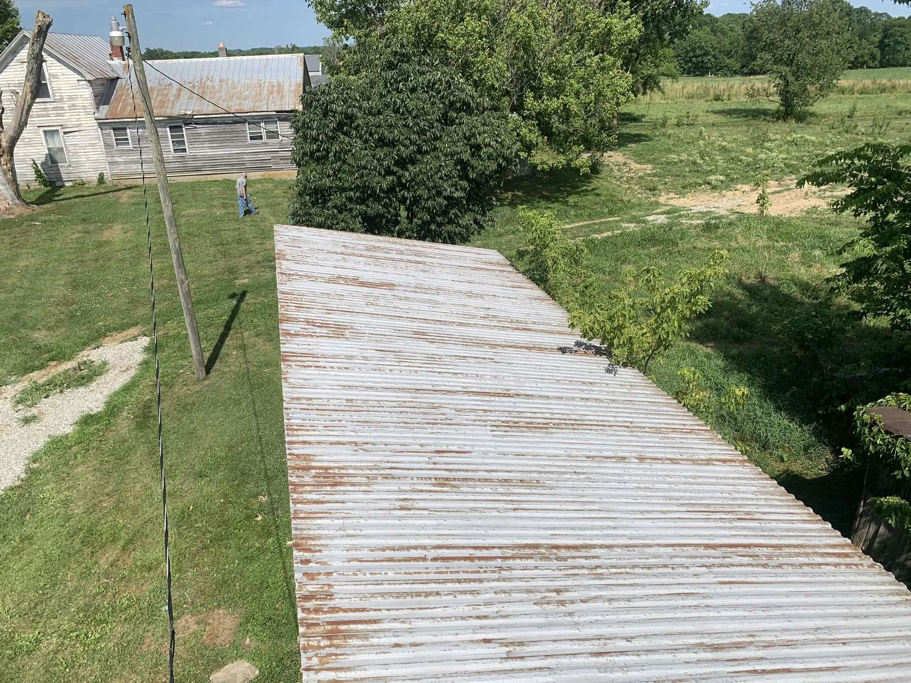 A rusted corrugated metal roof with green trees and a grassy yard in the background, and a person walking past an old wooden building.