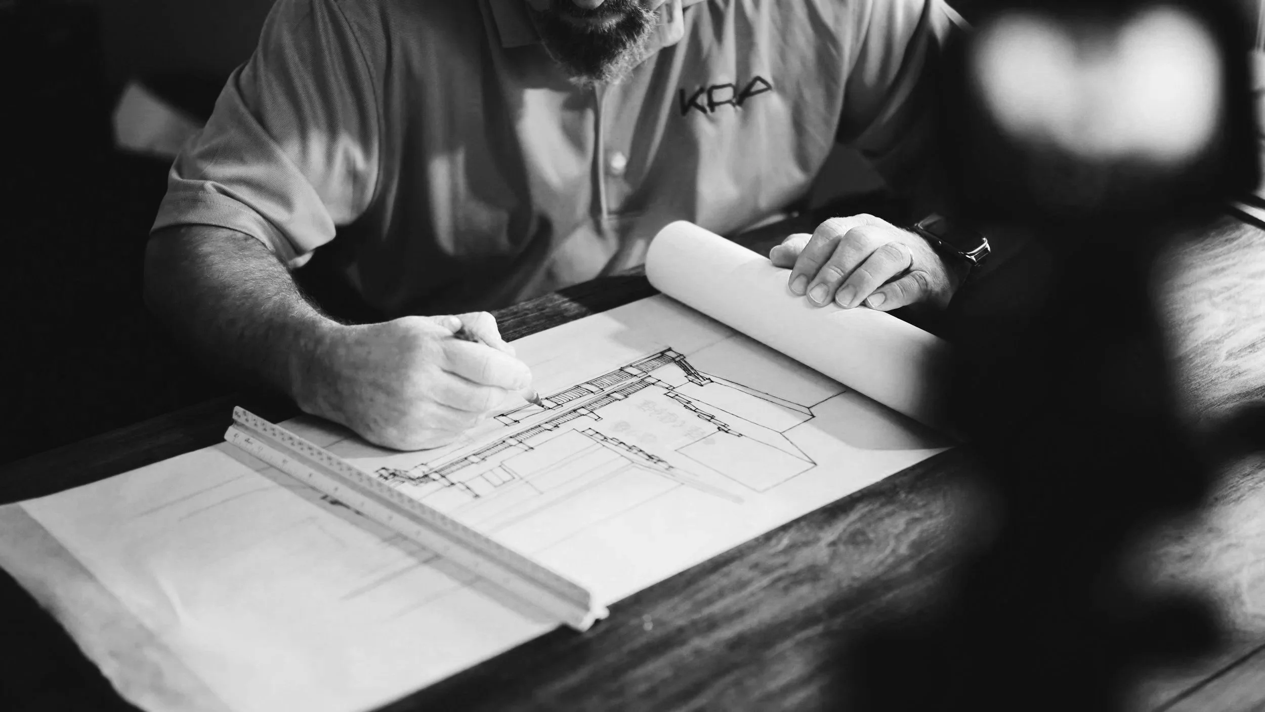 A person with a beard and wearing a shirt with 'KFA' on it is sitting at a wooden table, working on architectural blueprints, using a pencil and a ruler, with a measuring tape on the table.