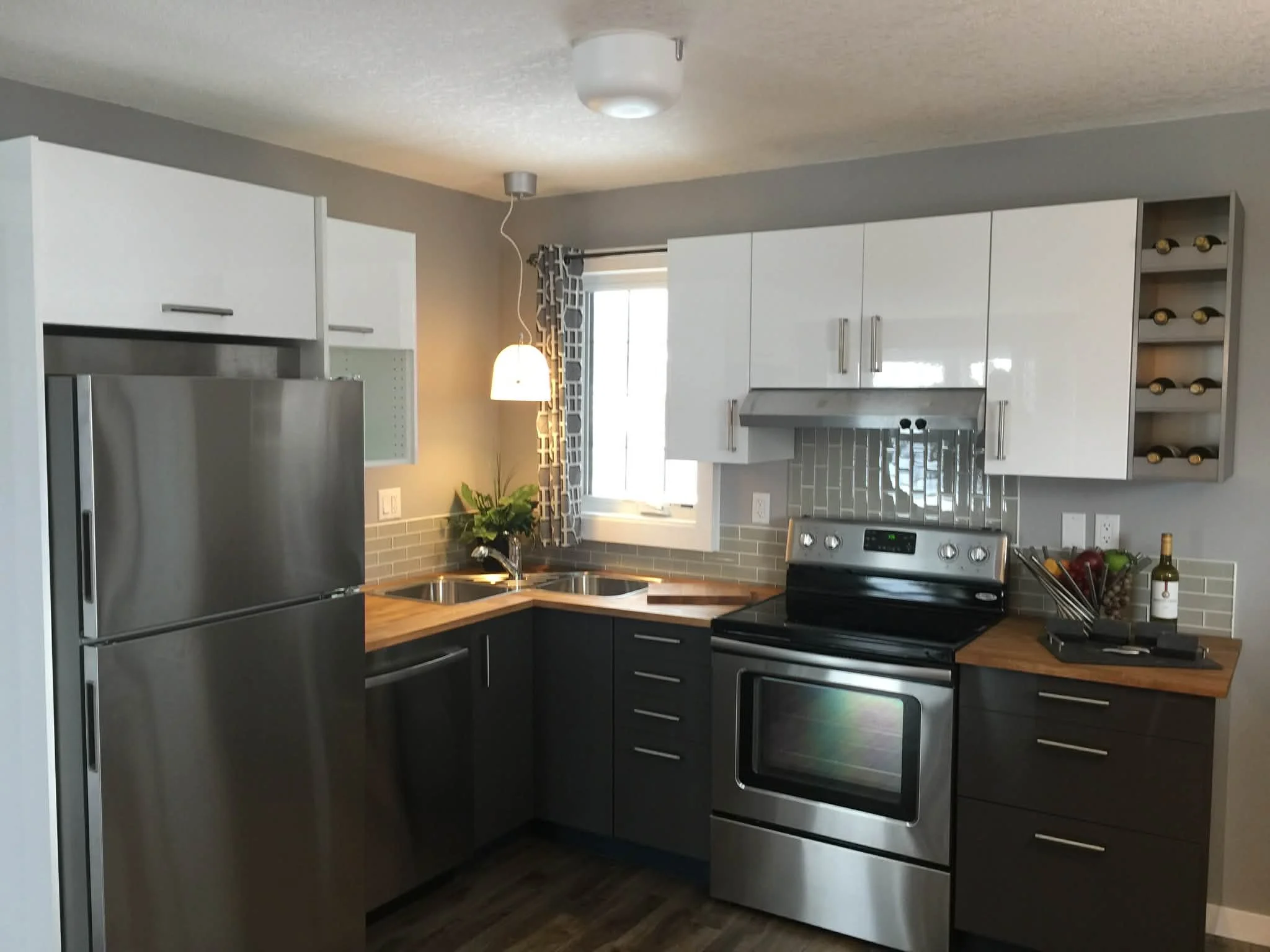 Modern kitchen with stainless steel refrigerator, black lower cabinets, white upper cabinets, gray subway tile backsplash, window with patterned curtains, pendant light, and various kitchen items.