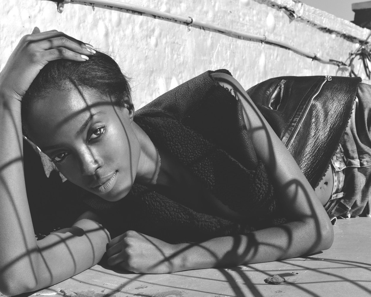 Black and white photo of a woman with short hair lying on a wooden surface, resting her head on her hand, looking directly at the camera. She is wearing a sleeveless top and a leather jacket.
