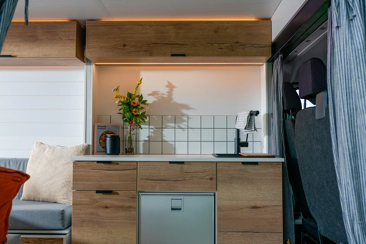 A small kitchenette with wooden cabinets, a black faucet, a white tiled backsplash, a framed picture, a black container, a vase with flowers, a gray sofa with a beige pillow, and a refrigerator beneath the countertop.