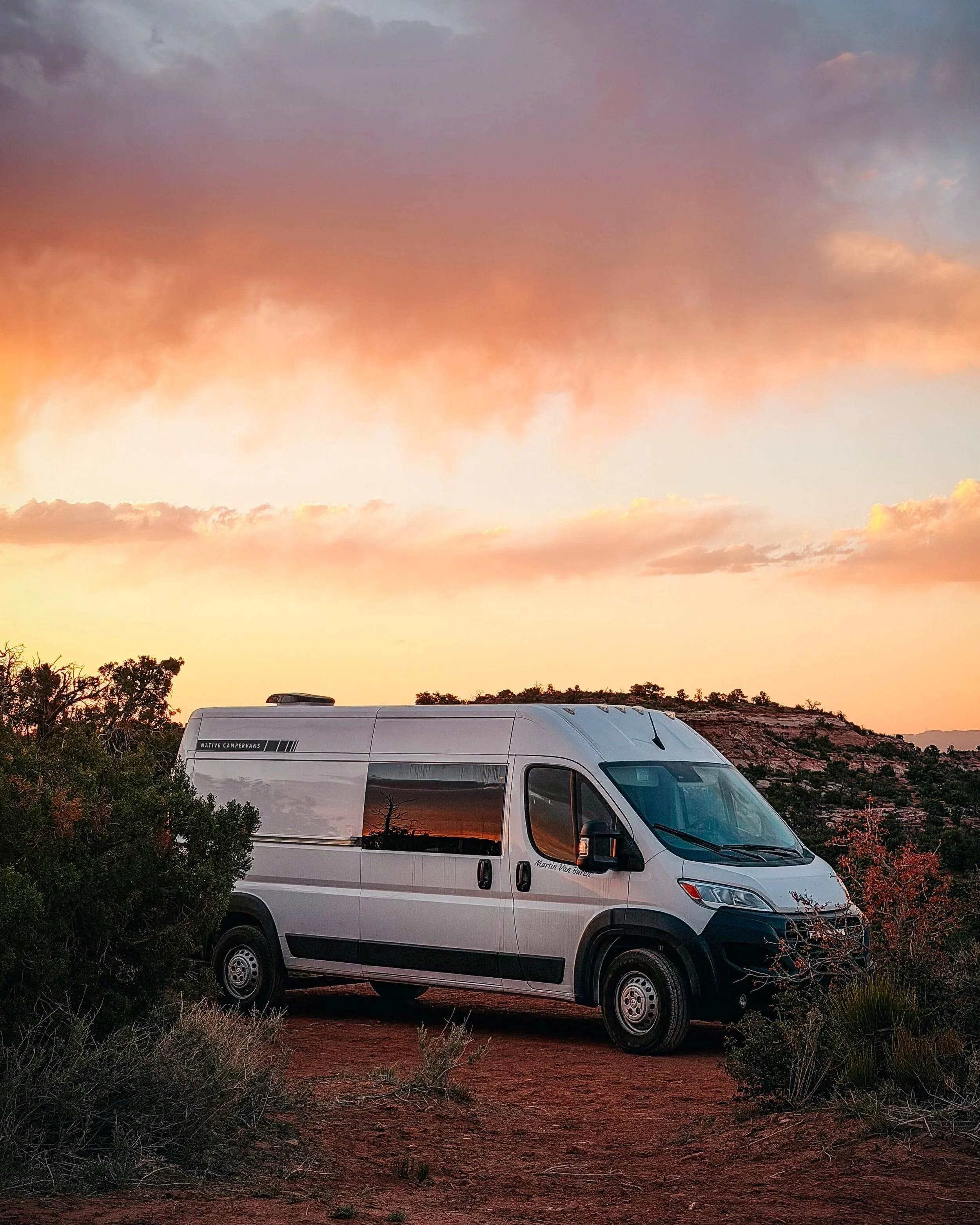 A white camper van parked on a dirt road with bushes and trees in the foreground, a rocky hillside in the background, and a colorful sunset sky with clouds.