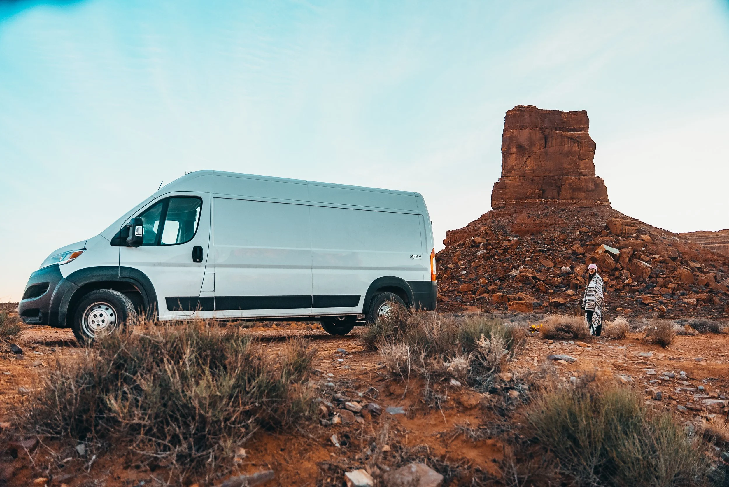 A white van parked on a dirt area in a desert landscape with a large red rock formation in the background and a person standing nearby.