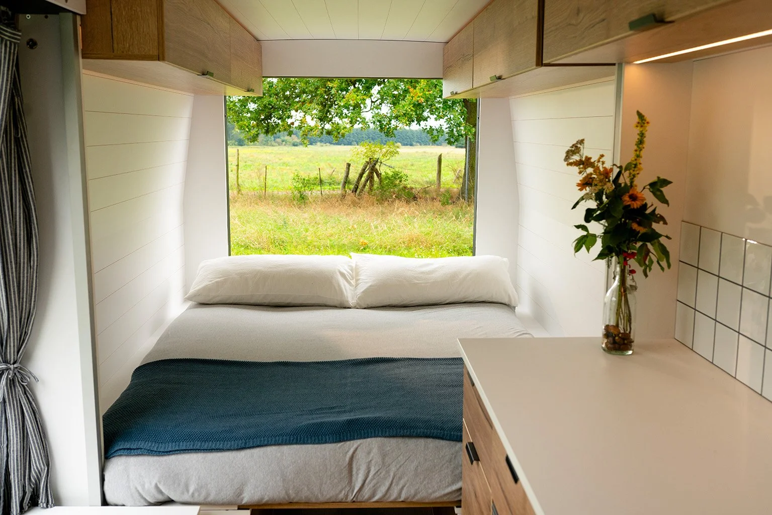 Inside view of a cozy bedroom with a large window overlooking a green field and trees, a bed with white bedding, and a nightstand with a vase of flowers.