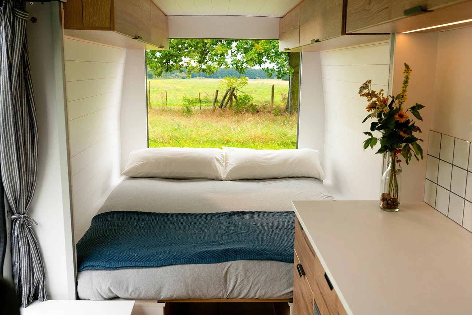 Room with a bed and a large window overlooking green fields and trees outside.