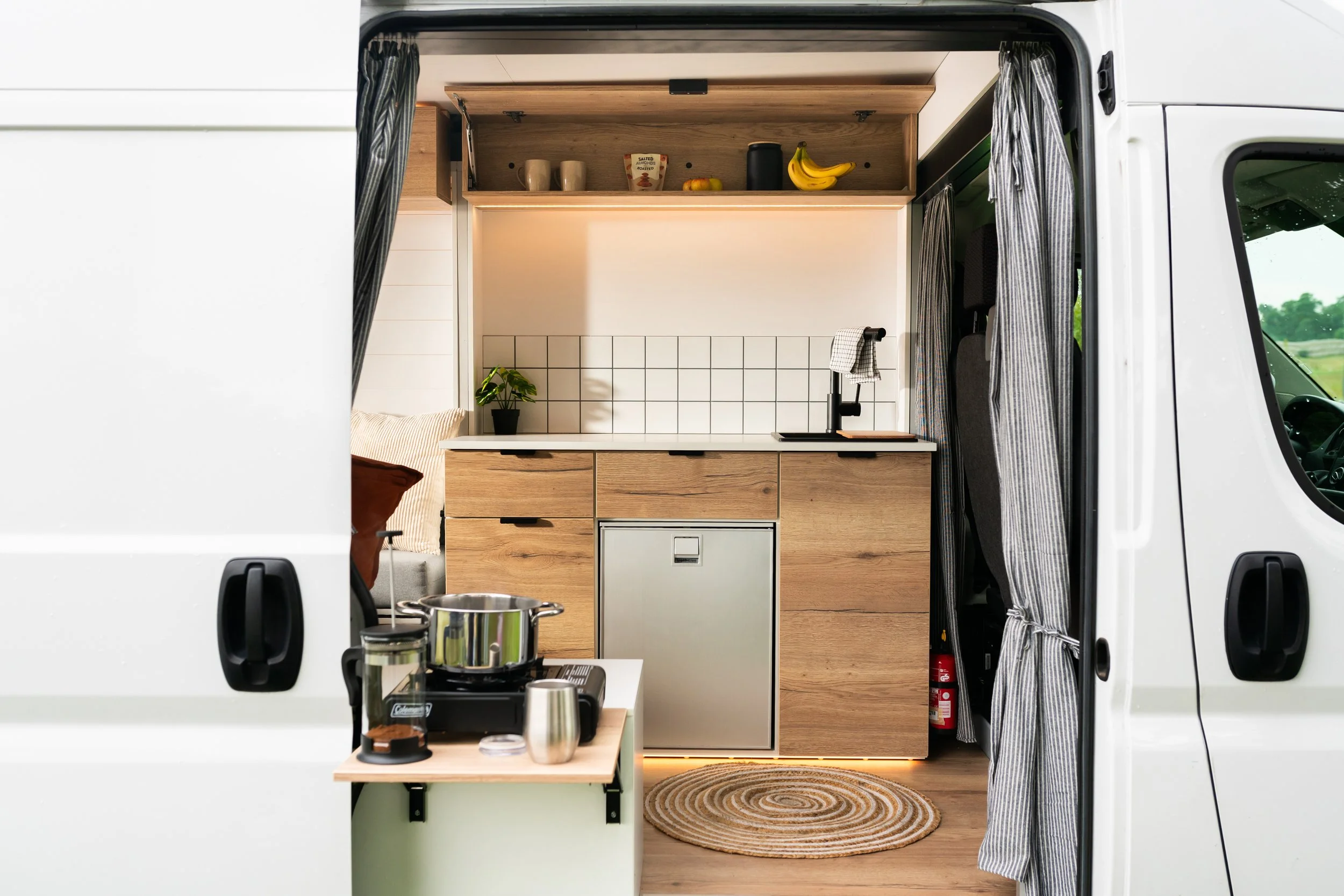 Interior of a compact camper van showing a wooden kitchen area with a small fridge, a sink with a black faucet, a potted plant, a stove with pots and pans, and a seating area with cushions. The van has curtains on the sides and windows showing greenery outside.
