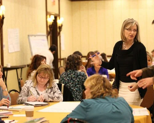 A woman standing and speaking to a group of seated women in a conference room.