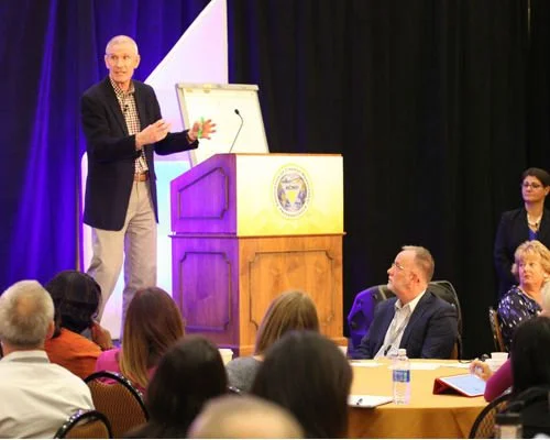 A speaker at a conference or seminar addressing an audience, standing next to a podium with a seal, with a black curtain backdrop.