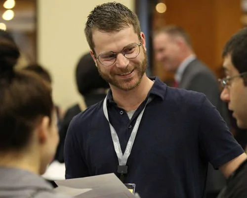 A smiling man with glasses and a beard talking to a group of people at a conference or social event.