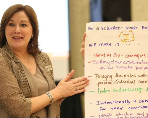 A woman with shoulder-length brown hair, wearing a beige shirt and blazer, standing or sitting by a window. She is pointing to a whiteboard with handwritten notes in various colors.