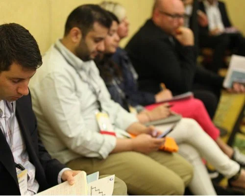 People seated in a row at a conference or seminar, reading papers or using tablets
