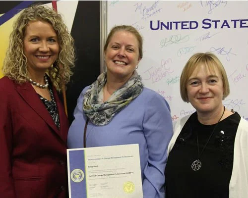 Three women standing together, with one holding a certificate, in front of a whiteboard with signatures, and a sign that says "United States".