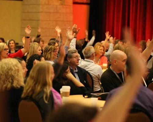 Audience members at a conference raising their hands during a vote or discussion.