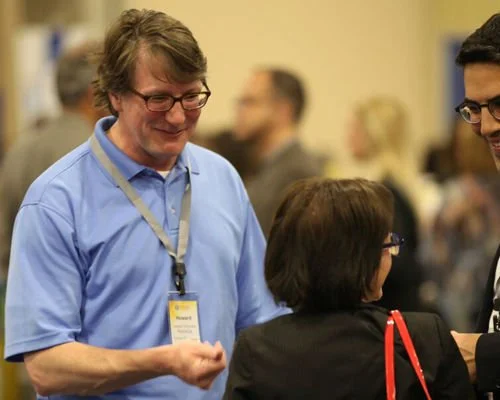 Three people engaged in conversation at a professional event, with name badges, in an indoor setting.