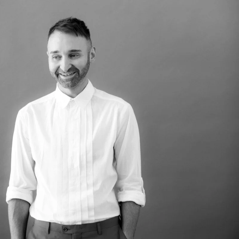 Black and white photo of a smiling man with a beard, wearing a white shirt with the sleeves rolled up, standing against a plain background.