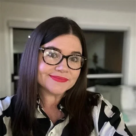 A woman with long dark hair and glasses, smiling in an indoor setting.