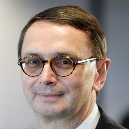 Close-up of a man with short brown hair and glasses, wearing a suit, smiling subtly.